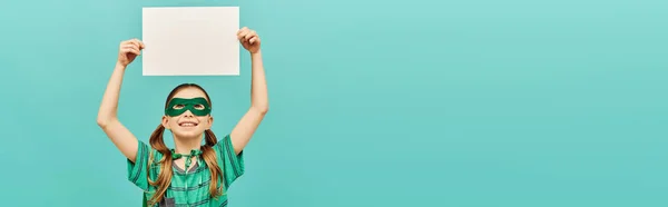 Glückliches Frühchen in grüner Superheldenmaske mit leerem Papier über dem Kopf und Blick nach oben auf blauem Hintergrund, Konzept zum Weltkinderschutztag, Banner — Stockfoto
