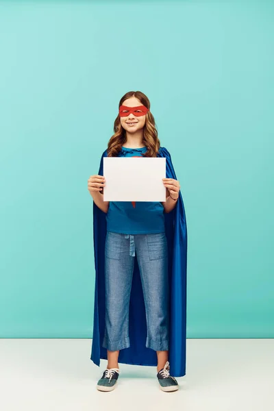 Позитивна дівчина в костюмі супергероя з плащем і червоною маскою, що стоїть з чистим папером і дивиться на камеру, святкуючи день захисту дітей на синьому фоні — стокове фото