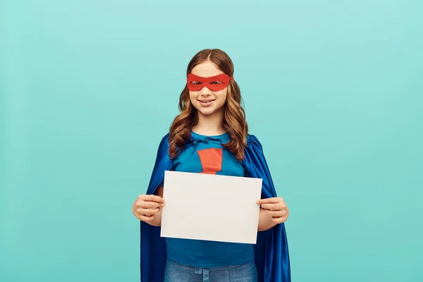Menina pré-adolescente positiva em traje de super-herói com máscara vermelha em pé com papel em branco e olhando para a câmera no fundo azul, conceito de dia de proteção da criança mundial — Fotografia de Stock