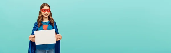 Garoto pré-adolescente positivo em traje de super-herói com capa e máscara vermelha em pé com papel em branco e olhando para a câmera no fundo azul, conceito de dia feliz das crianças, banner — Fotografia de Stock