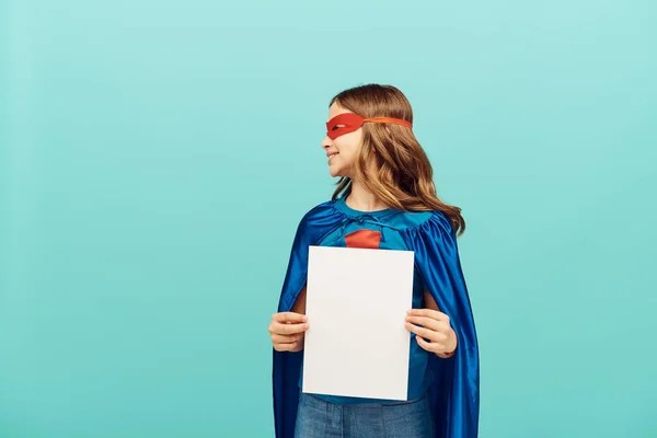 Беззаботная девочка-подросток в костюме супергероя с красной маской, стоящей с чистой бумагой и глядя в сторону на синий фон, концепция Всемирного дня защиты детей — стоковое фото