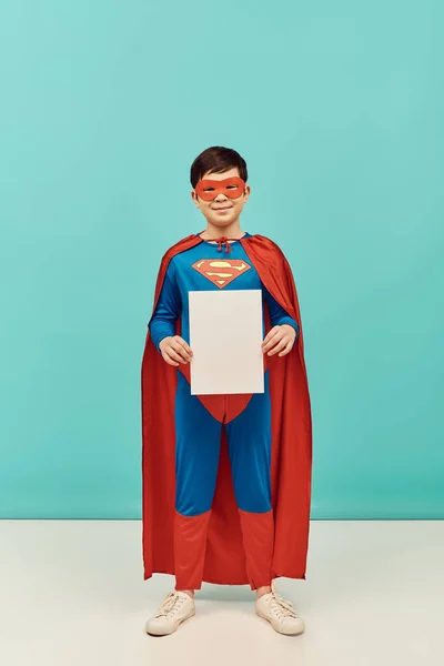 Full length of preteen asian boy in superhero costume with mask and cloak standing with blank paper and looking at camera on blue background, World child protection day concept — Stock Photo