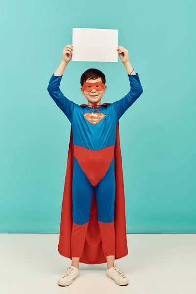 Full length of preteen asian boy in superhero costume with mask and cloak holding blank paper above head and looking at camera on blue background, International Child Protection Day concept — Stock Photo