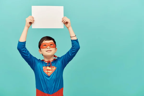 Frühchen asiatischer Junge im Superheldenkostüm mit Maske, leeres Papier über dem Kopf und Blick nach oben auf blauem Hintergrund, Konzept zum Internationalen Kinderschutztag — Stockfoto
