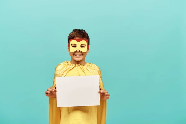 Улыбающийся многорасовый мальчик в желтом костюме супергероя с маской, держа чистую бумагу на синем фоне, счастливый день ребенка концепции — стоковое фото