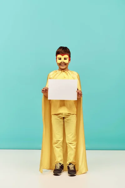 Longitud completa de niño multirracial positivo en traje de superhéroe amarillo con máscara sosteniendo papel en blanco sobre fondo azul, concepto del Día Internacional de la Protección del Niño - foto de stock