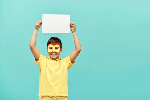 Niño multirracial sonriente en traje de superhéroe amarillo con máscara sosteniendo papel en blanco sobre la cabeza sobre fondo azul, concepto del día internacional de los niños - foto de stock