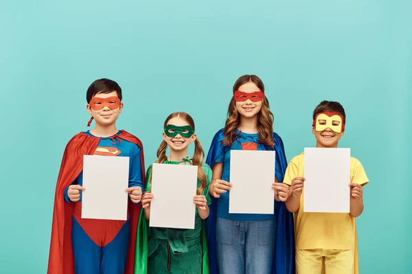 Щасливі міжрасові діти в барвистих костюмах супергероя з масками посміхаються і тримають чисті папери, дивлячись на камеру на синьому тлі в студії, концепцію Дня захисту дітей — стокове фото