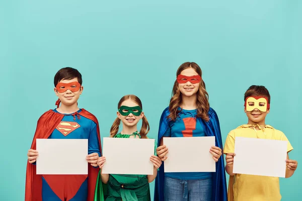 Усміхнені міжрасові діти в барвистих костюмах супергероїв з масками, що тримають порожні папери, дивлячись на камеру на синьому фоні в студії, концепція щасливого дня дітей — стокове фото