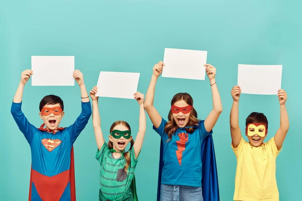 Щасливі міжрасові діти в барвистих супергеройських костюмах з масками, що тримають чисті папери над головами, дивлячись на камеру на синьому фоні в студії, концепція Всесвітнього дня захисту дітей — стокове фото