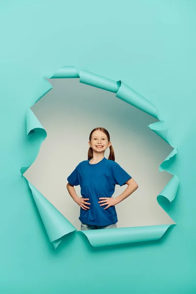 Счастливая рыжая девочка-подросток в футболке смотрит в камеру, позируя руками на бедрах вокруг синей рваной бумажной дыры на белом фоне, концепция Международного дня защиты детей — стоковое фото