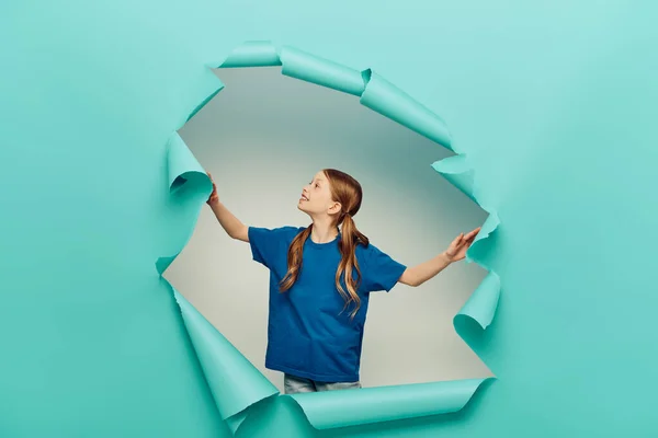 Menina ruiva alegre em t-shirt sorrindo e olhando para buraco de papel rasgado azul no fundo branco, conceito do Dia Internacional da Proteção à Criança — Fotografia de Stock