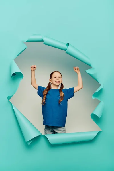 Взволнованная и рыжая девочка-подросток в футболке, смотрящая на камеру, показывая жест силы вокруг синей рваной бумажной дыры на белом фоне, концепция Международного дня защиты детей — стоковое фото