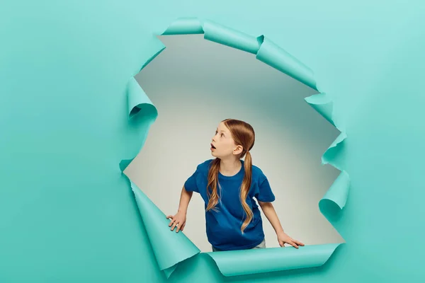 Шокированная рыжая девочка-подросток в футболке, смотрящая на голубую порванную бумагу, создающую дыру на белом фоне, концепция Международного дня защиты детей — стоковое фото