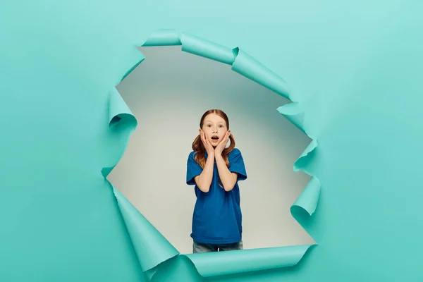 Scared redhead preteen girl in t-shirt looking at camera while touching face with hands around blue torn paper hole on white background, Global Children Day concept — Stock Photo