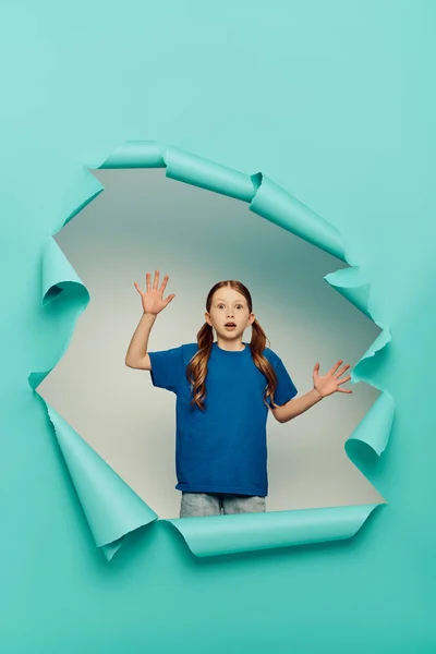 Verängstigtes rothaariges Mädchen, das in die Kamera schaut, während es gestikuliert, in der Nähe eines blauen zerrissenen Papierlochs auf weißem Hintergrund, Konzept für den Internationalen Kindertag — Stockfoto