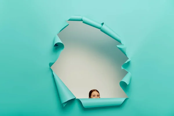 Shocked redhead preteen girl looking at camera through blue torn paper hole on white background, International Child Protection Day concept — Stock Photo