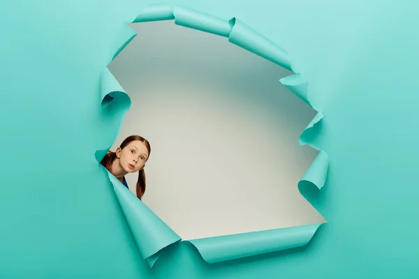 Surprised redhead preteen girl looking at camera through blue torn paper hole on white background, International Child Protection Day concept — Stock Photo