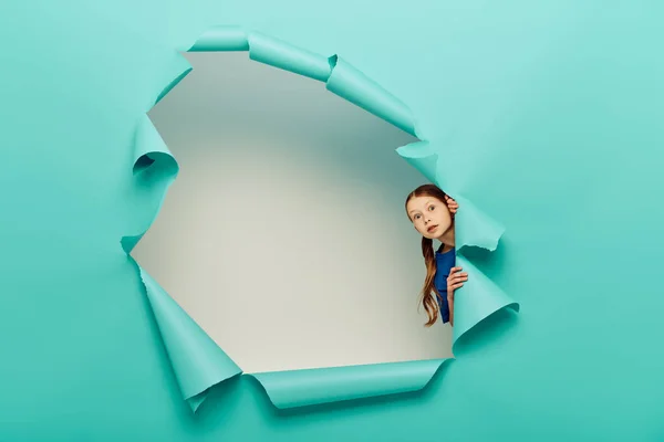 Scared red haired preteen girl in blue t-shirt looking at camera while standing behind hole in blue paper on white background background — Stock Photo