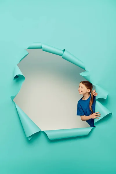 Cheerful red haired girl in t-shirt looking away while touching hole in blue paper and celebrating International Day for Protection of Children on white background — Stock Photo