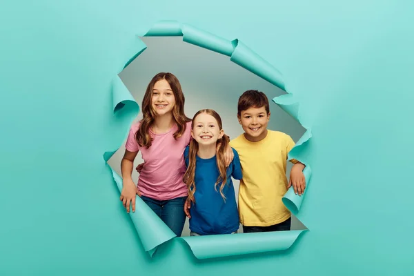 Позитивные межрасовые дети в разноцветных футболках, смотрящие в камеру во время празднования Дня защиты детей и стоящие за дырой на синем бумажном фоне — стоковое фото