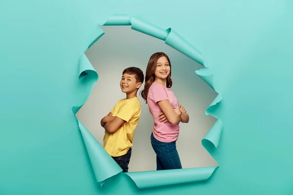Smiling interracial preteen kids in colorful t-shirts crossing arms while standing back to back and celebrating child protection day behind hole in blue paper background — Stock Photo