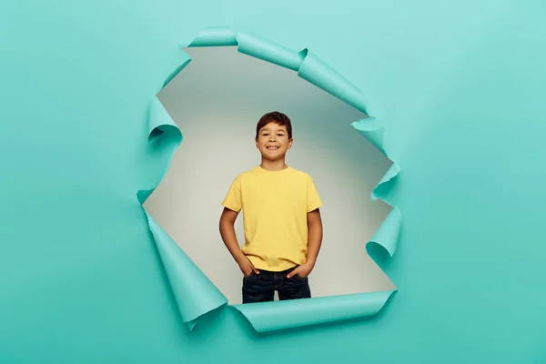 Smiling multicultural boy holding hands in pockets of pants and looking at camera while celebrating child protection day behind hole in blue paper background — Stock Photo