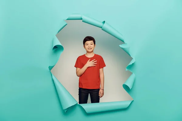 Sonriente asiático preadolescente chico en rojo camiseta mirando a la cámara durante la celebración del día de la protección del niño y de pie detrás del agujero en fondo de papel azul - foto de stock