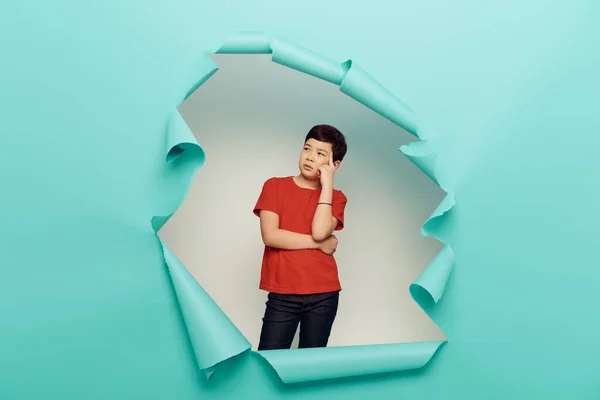Pensive asian preteen boy in red t-shirt looking away while standing behind hole in blue paper on white background, World Child protection day concept — Stock Photo