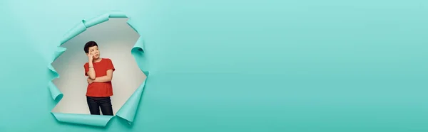 Nachdenklicher asiatischer Bub schaut weg, während er hinter einem Loch in blauem Papier auf weißem Hintergrund steht, Konzept zum Weltkinderschutztag, Banner — Stockfoto