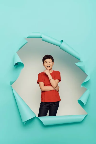 Feliz niño preadolescente asiático en camiseta roja mirando a la cámara durante la celebración del Día Mundial de la Protección del Niño detrás del agujero en papel azul sobre fondo blanco - foto de stock
