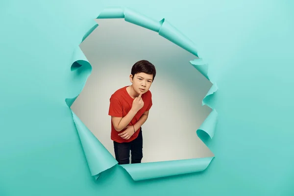 Pensive asiatische Preteen Boy in rotem T-Shirt Blick in die Kamera während der internationalen Kindertagsfeier, während hinter Loch in blauem Papier Hintergrund stehen — Stockfoto