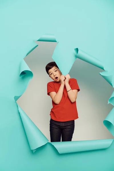 Shocked asiático preadolescente chico rojo camiseta mirando cámara y tocando agujero en azul papel mientras celebrando el día de la protección del niño en blanco fondo - foto de stock