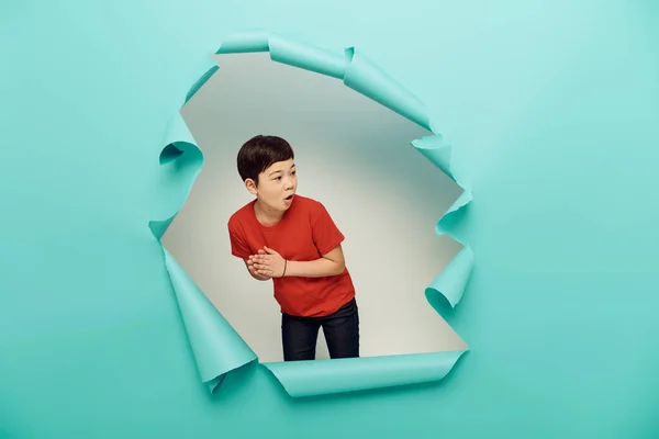 Surprised asian preteen boy in casual red t-shirt looking away during child protection day celebration behind hole in blue paper on white background — Stock Photo