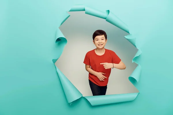 Positivo asiático preadolescente chico en rojo camiseta apuntando con el dedo durante la protección del niño día celebración detrás de agujero en azul papel y en blanco fondo - foto de stock
