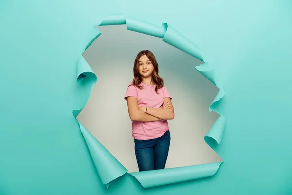 Lächelndes Mädchen in legerer Kleidung, das die Arme verschränkt und während der Kinderschutztagsfeier hinter einem Loch im blauen Papierhintergrund in die Kamera blickt — Stockfoto