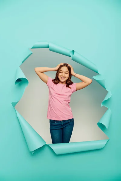 Позитивна дівчина в рожевій футболці, що торкається волосся, стоїть за отвором у синьому папері на білому тлі, концепція щасливого дня дітей — стокове фото