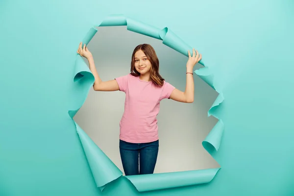Carefree preteen girl in casual clothes looking at camera during child protection day celebration behind hole in blue paper on white background — Stock Photo
