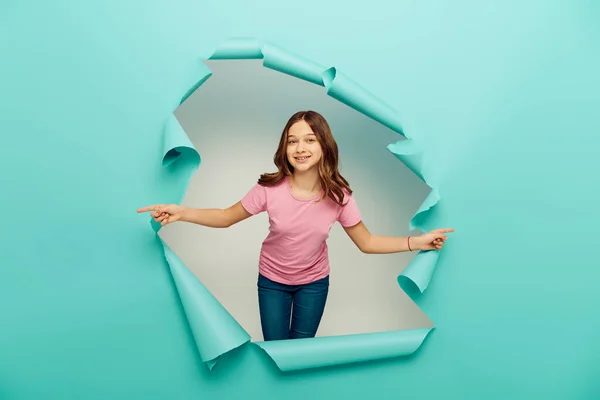 Joyful preteen girl in casual clothes pointing with fingers during international children day celebration behind hole in blue paper background — Stock Photo