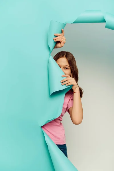 Ребенок-подросток в повседневной одежде, закрывающий лицо и смотрящий на камеру сквозь дыру в голубой бумаге, скрываясь на сером фоне, концепция Международного дня защиты детей — стоковое фото