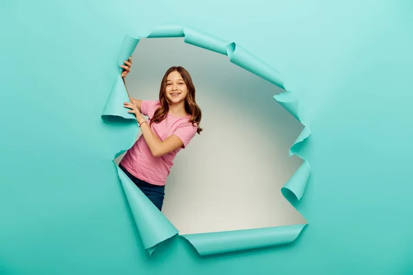 Cheerful preteen girl in casual clothes having fun while celebrating child protection day and standing behind hole in blue paper on white background — Stock Photo