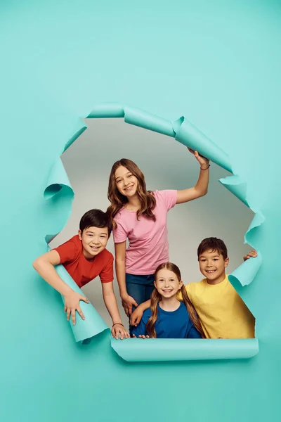Alegre grupo de niños multiétnicos en ropa colorida mirando a la cámara durante el día de protección infantil mientras están de pie detrás del agujero en el fondo de papel azul - foto de stock