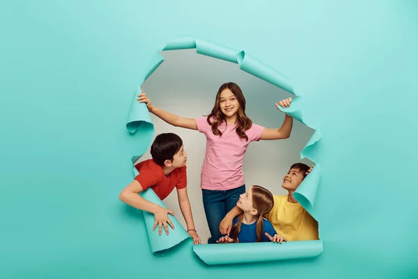 Smiling interracial kids in colorful clothes looking at friend while celebrating international children day behind hole in blue paper background — Stock Photo