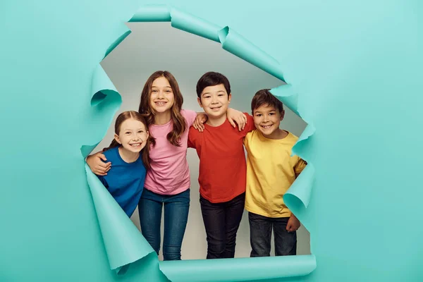Niños preadolescentes multiétnicos sonrientes en camisetas coloridas abrazándose durante la celebración del día de protección de los niños detrás del agujero en el fondo de papel azul - foto de stock
