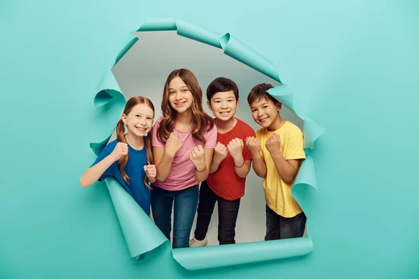 Smiling multiethnic group of kids looking at camera and showing fists during child protection day celebration behind hole in blue paper background — Stock Photo
