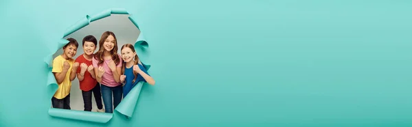 Enfants multiethniques joyeux dans des vêtements colorés montrant poings pendant la journée internationale des enfants tout en se tenant derrière le trou dans fond de papier bleu, bannière — Photo de stock