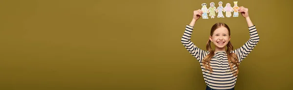 Carefree preteen menina de cabelos vermelhos em camisa listrada segurando personagens de papel desenhado durante a celebração do dia de proteção infantil no fundo cáqui com espaço de cópia, banner — Fotografia de Stock