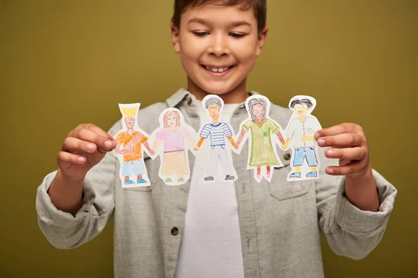 Blurred and positive multiracial boy holding drawn paper characters during international child protection day celebration on khaki background — Stock Photo