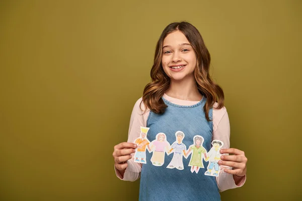 Smiling preteen girl in denim sundress holding drawn paper characters and looking at camera during child protection day celebration on khaki background — Stock Photo