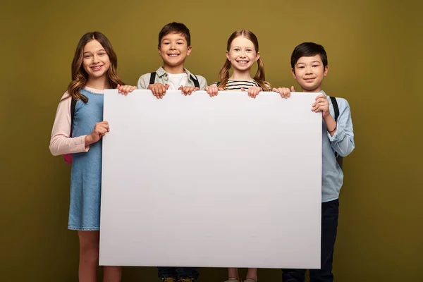 Lächelnde Kinder mit Rucksäcken mit leeren Plakaten und Blick in die Kamera bei der Kinderschutztagsfeier auf khakifarbenem Hintergrund — Stockfoto